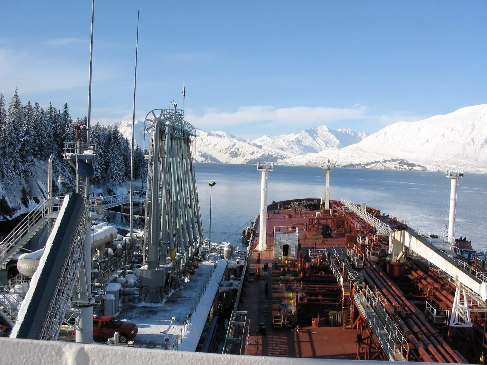 Tanker deck with red cargo piping alongside snow-covered loading arms at the Valdez terminal, with a tug at the bow and snowy mountains across the water.